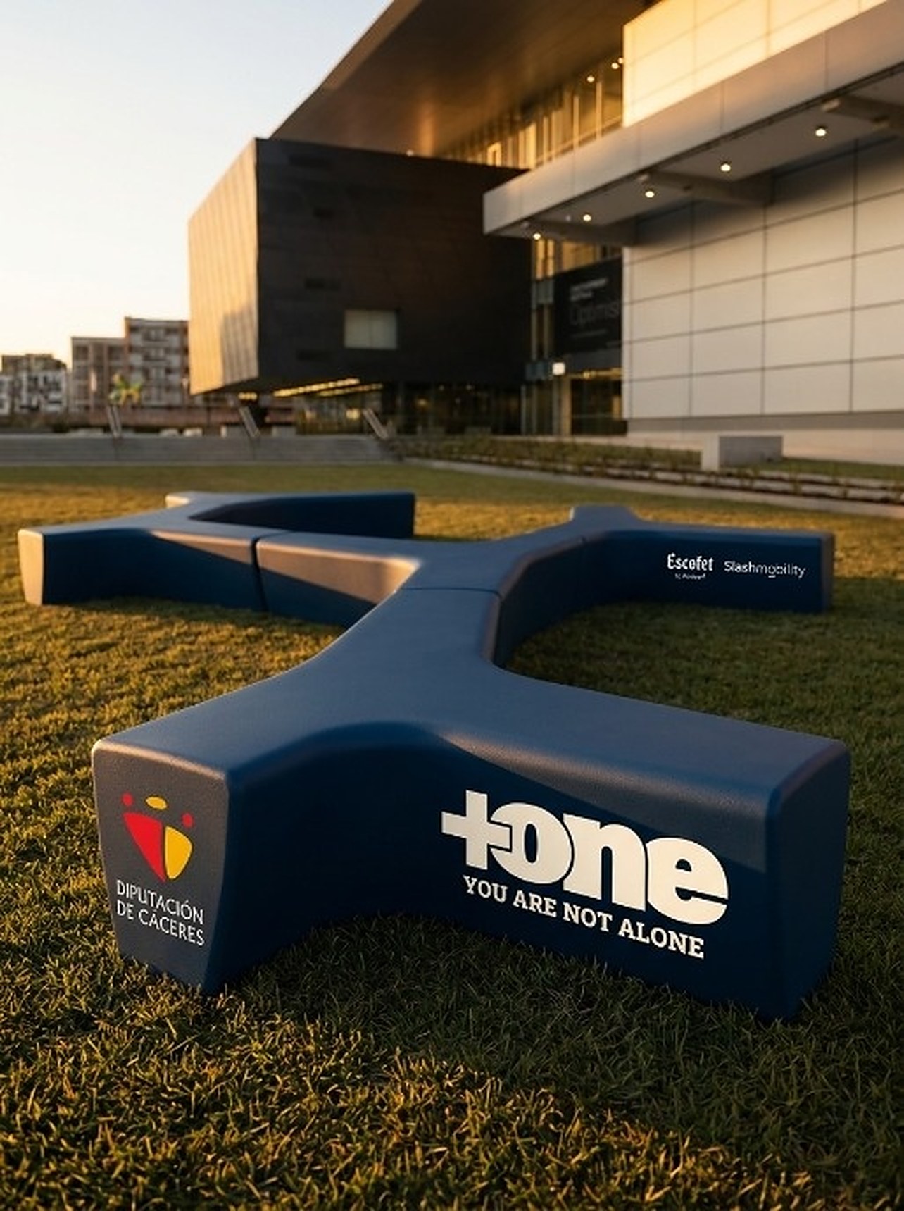 Banco de la Amistad de +one, en forma de Y, sobre el césped al atardecer frente a un edificio.